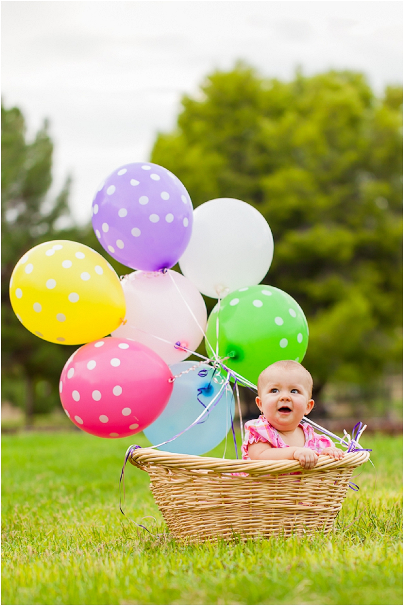 A one year old girl sitting in a basket with balloons tied to it. 