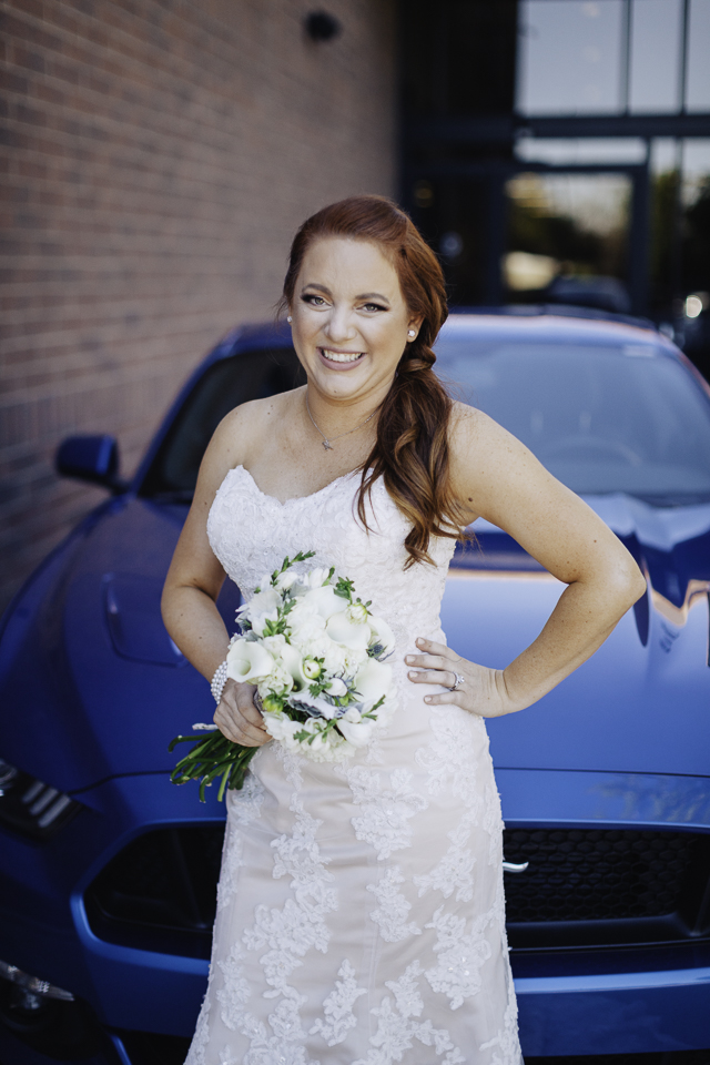Bride posing in front of groom's blue Mustang sports car. 