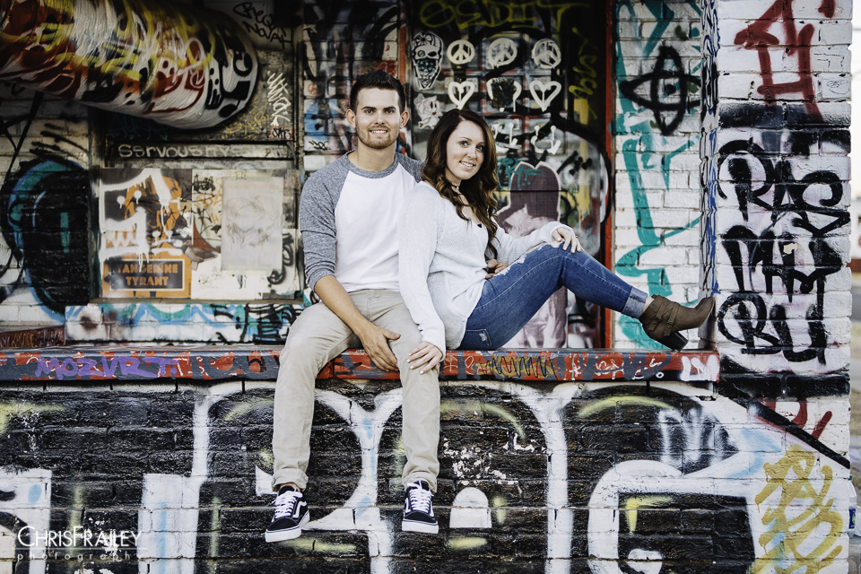 Engaged couple sitting outside a graffiti sprayed painted house.