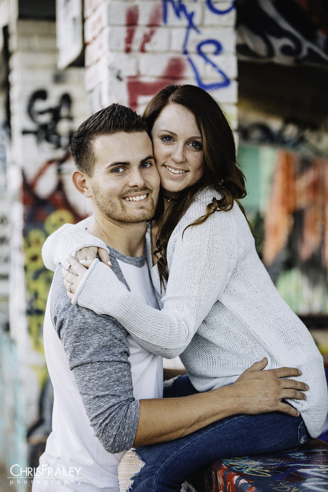 Engaged couple holding each other on porch of a graffiti spray painted house. 