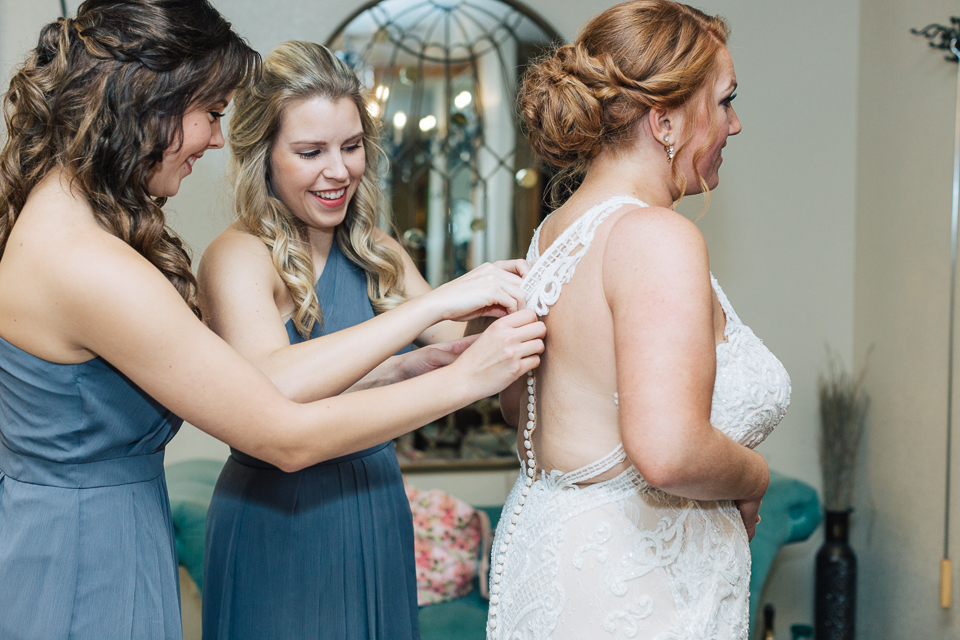 Twin sisters of the bride help her with button up the wedding dress.