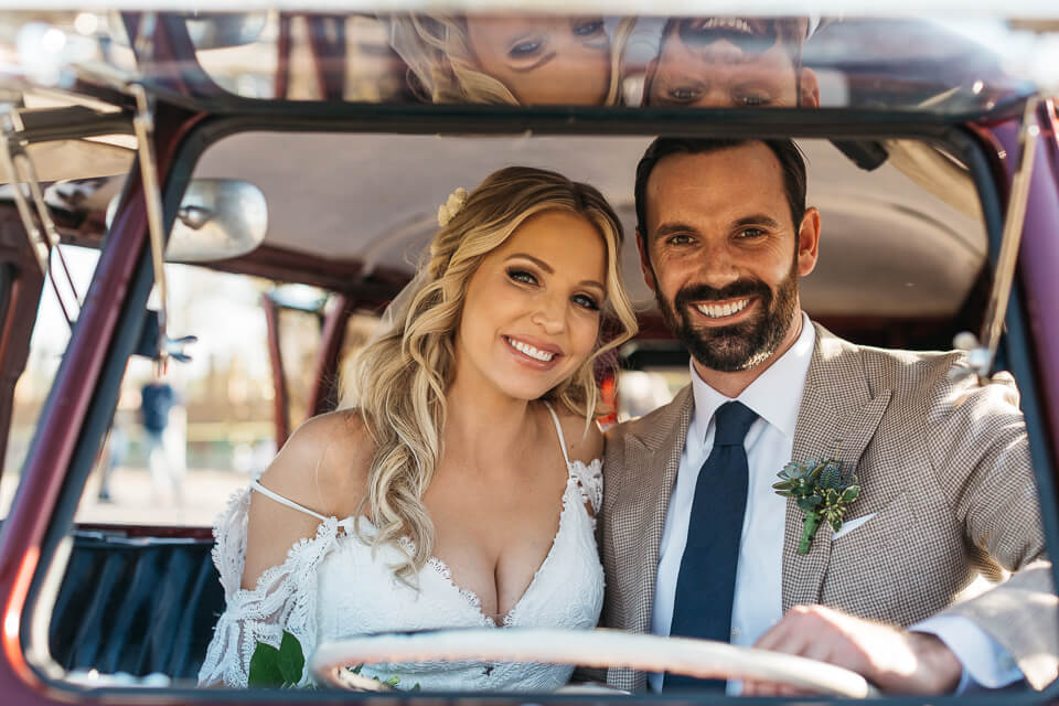 Bride and groom sitting in an old VW bus with windshield open.