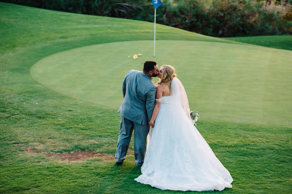 Bride and groom kissing while standing next to a golf green. 
