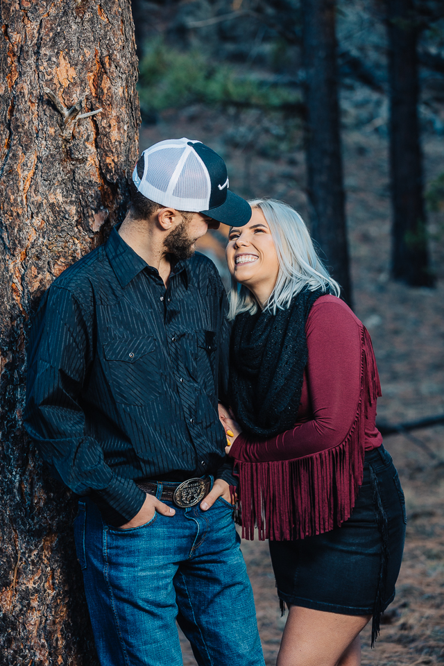 Couple leaning against a pine tree smiling at each other.