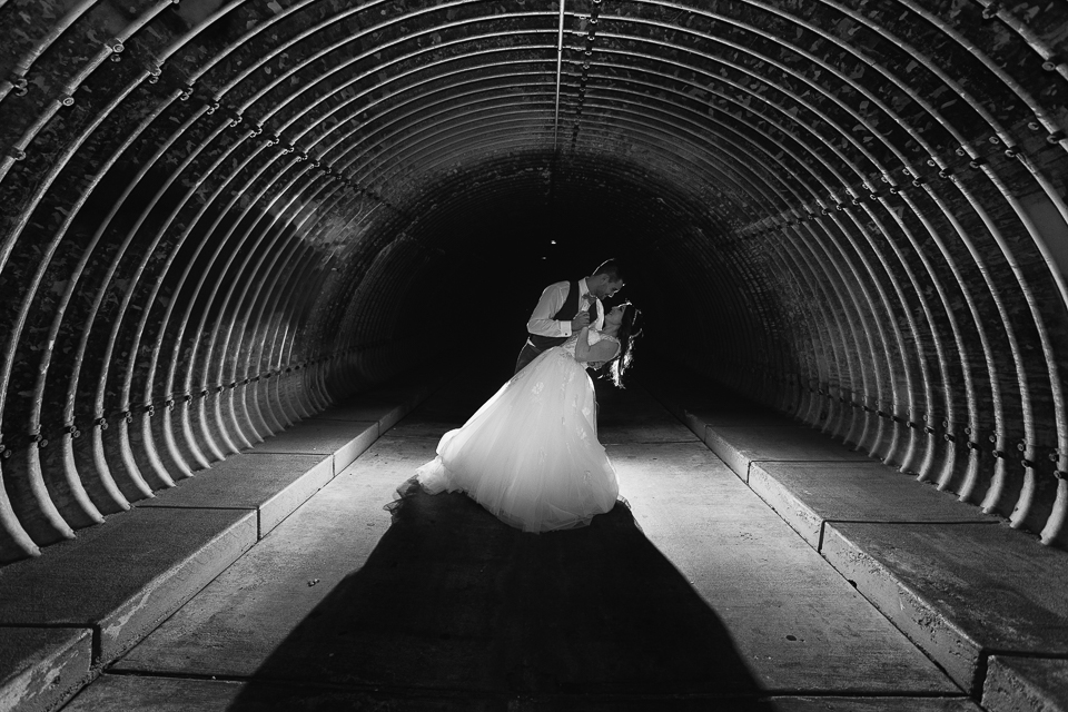 Groom dipping his bride inside a large metal tunnel.