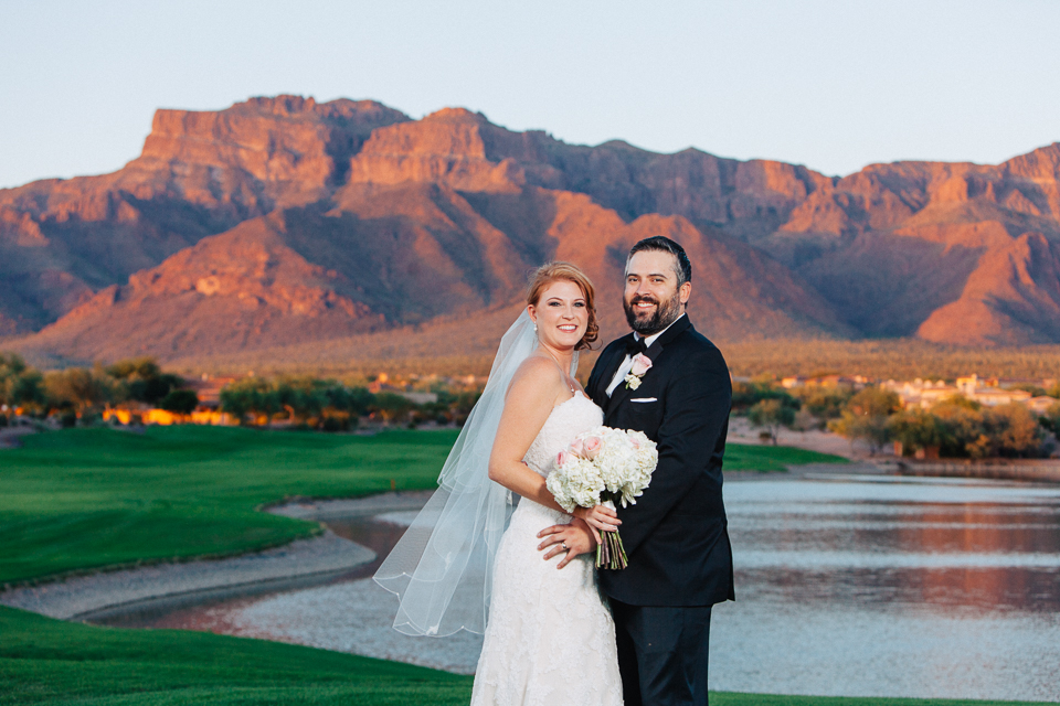 Bride and groom posing by lake with Superstition Mountains in the background.